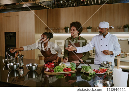 African American female intern helping her Indian master with kitchenware while her mate writing down recipe African American female intern helping her Indian master with kitchenware while her mate writing down recipe 118946631