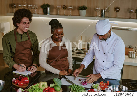 High angle shot of chef in white uniform cutting red onion while his biracial interns writing down his actions 118946648