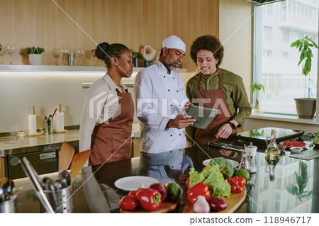 Professional chef holding touchpad with new menu and discussing it with his assistants while they standing in center of kitchen Professional chef holding touchpad with new menu and discussing it with his assistants while they standing in center of kitchen 118946717