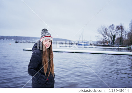 beautiful young girl with long blond hair in a hat and a long down jacket on the pier of a winter lake 118948644