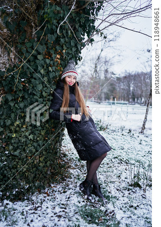 a beautiful young girl with long blond hair in a hat and a long down jacket plays with snow and enjoys winter nature a beautiful young girl with long blond hair in a hat and a long down jacket plays with snow and enjoys winter nature 118948661