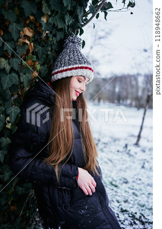 a beautiful young girl with long blond hair in a hat and a long down jacket plays with snow and enjoys winter nature a beautiful young girl with long blond hair in a hat and a long down jacket plays with snow and enjoys winter nature 118948662