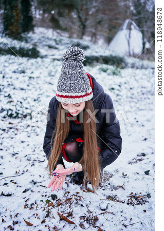 a beautiful young girl with long blond hair in a hat and a long down jacket plays with snow and enjoys winter nature a beautiful young girl with long blond hair in a hat and a long down jacket plays with snow and enjoys winter nature 118948678