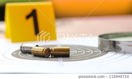 Empty bullet casings on a shooting target background, with blur black number 1 on yellow label and magnifying glass placed nearby, blending the themes of crime investigation. 118948720