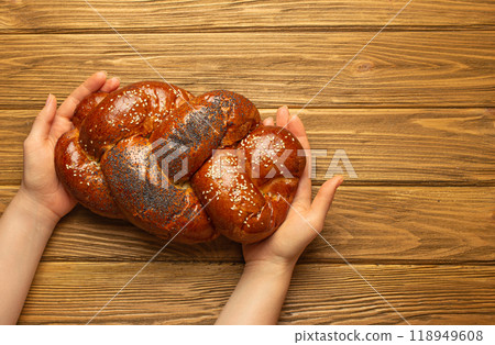 Female hands holding freshly baked Challah bread covered with poppy and sesame seeds, top view on rustic wooden background, traditional festive Jewish cuisine Female hands holding freshly baked Challah bread covered with poppy and sesame seeds, top view on rustic wooden background, traditional festive Jewish cuisine 118949608