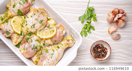 Raw uncooked chicken legs marinated with seasonings, herbs, lemon in white ceramic casserole top view on light wooden rustic background. Preparing healthy meal with marinated chicken drumsticks 118949610