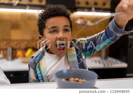 Dark-skinned boy sitting at the table and eating cereals Dark-skinned boy sitting at the table and eating cereals 118949695