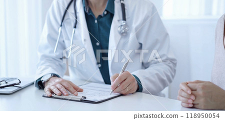 Doctor and a patient. The physician, wearing a white medical coat over a green shirt, is filling out a medical record form during a consultation in the clinic. Medical service 118950054