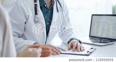 Doctor and a patient. The physician, wearing a white medical coat over a green shirt, is gesturing with his hands during a consultation in the clinic. Medicine 118950059