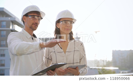 Two architects colleagues, wearing safety helmets and glasses, are standing near each other and discussing a construction site progress during sunrise or sunset. Architecture and engineering 118950066