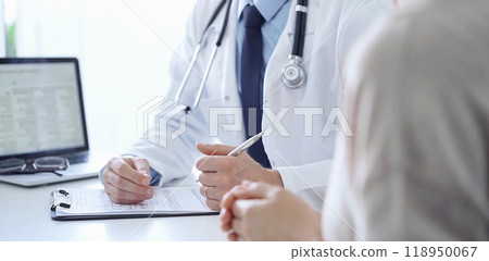 A doctor and a patient. The physician, wearing a white medical coat over a blue shirt and tie, is filling out a medical record form during a consultation in the clinic. Medical service 118950067