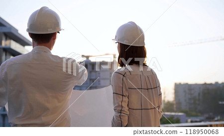 Two architects, wearing hard hat and safety glasses, are examining blueprints at a construction site during sunrise or sunset, viewed from the back. Architecture and engineering 118950068