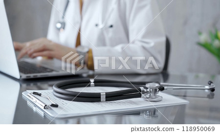 Stethoscope and clipboard with medication history records are on a glass desk behind a physician using a laptop computer. Medicine concept 118950069