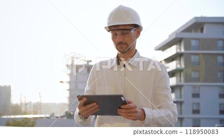 Man construction engineer or architect wearing white shirt and hard hat is using digital tablet while inspecting a building site at sunset, front view. Architecture and engineering concepts 118950083