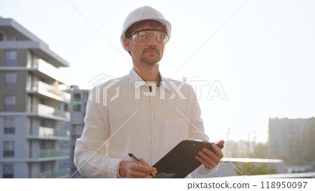Man construction engineer wearing white shirt and hard hat is making notes on a clipboard while inspecting a building site at sunset, front view. Architecture and engineering concepts 118950097