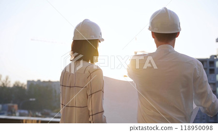Two architects, wearing hard hat and safety glasses, are examining blueprints at a construction site during sunrise or sunset, viewed from the back. Architecture and engineering 118950098