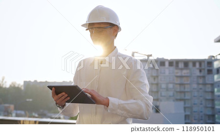 Man construction engineer or architect wearing white shirt and hard hat is using digital tablet while inspecting a building site at sunset, front view. Architecture and engineering concepts Man construction engineer or architect wearing white shirt and hard hat is using digital tablet while inspecting a building site at sunset, front view. Architecture and engineering concepts 118950101