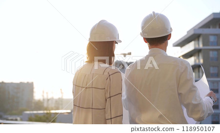 Man and woman architects constructor engineers wearing safety glasses and hardhats, are examining blueprints at a construction site during a sunset, back view. Architecture concept Man and woman architects constructor engineers wearing safety glasses and hardhats, are examining blueprints at a construction site during a sunset, back view. Architecture concept 118950107