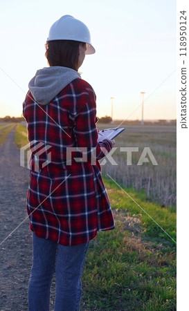 Woman engineer wearing a white protective helmet is taking notes with a clipboard in a field with wind turbines. Clean energy and engineering concept 118950124