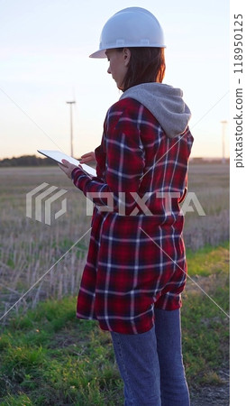 Woman engineer wearing a white protective helmet is taking notes with a clipboard in a field with wind turbines, as the sun sets. Clean energy and engineering concept Woman engineer wearing a white protective helmet is taking notes with a clipboard in a field with wind turbines, as the sun sets. Clean energy and engineering concept 118950125