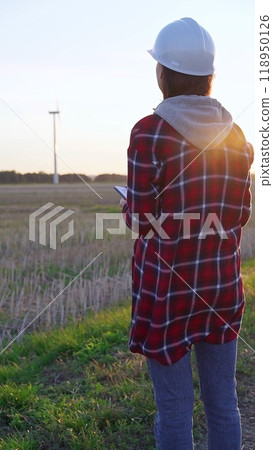 Woman engineer wearing a white protective helmet is taking notes with a clipboard in a field with wind turbines, as the sun sets. Clean energy and engineering concept 118950126