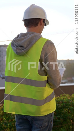 Male engineer with white helmet and safety vest is taking notes on a clipboard while inspecting a construction site at sunrise, vertical back view. Architecture and engineering Male engineer with white helmet and safety vest is taking notes on a clipboard while inspecting a construction site at sunrise, vertical back view. Architecture and engineering 118950151