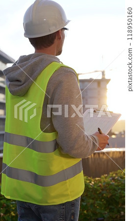 Male engineer with white helmet and safety vest is taking notes on a clipboard while inspecting a construction site at sunrise, vertical back view. Architecture and engineering 118950160