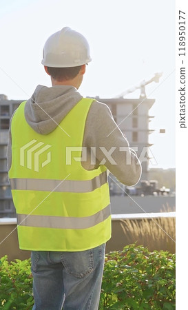 Man engineer with white helmet and safety vest is using tablet computer while inspecting a construction site at sunrise, back vertical view Man engineer with white helmet and safety vest is using tablet computer while inspecting a construction site at sunrise, back vertical view 118950177
