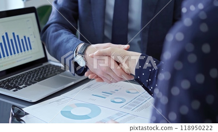 Two business people professionals shaking hands in office setting, sealing a deal with laptop and financial graphs visible. Business handshake over laptop and reports 118950222