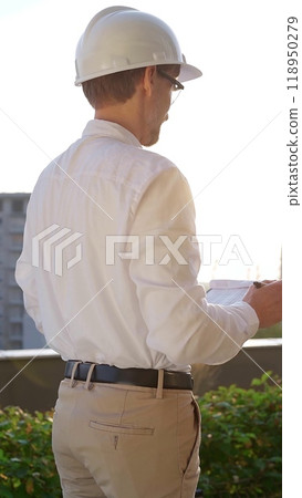 Man a construction engineer wearing white shirt and hard hat is making notes on a clipboard while inspecting a building site at sunset, back vertical view . Architecture concept Man a construction engineer wearing white shirt and hard hat is making notes on a clipboard while inspecting a building site at sunset, back vertical view . Architecture concept 118950279