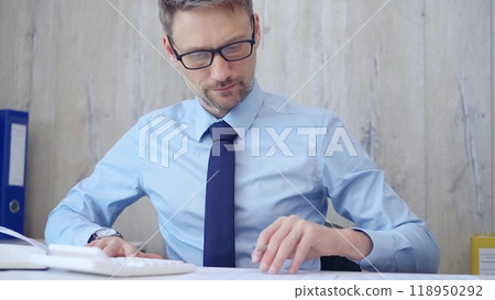 Male accountant with blue t-shirt concentrating while calculating costs and taking notes at his desk. Taxes, audit in business 118950292