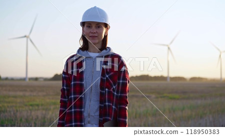 Female engineer is standing straight on a field with wind turbines, as the sun sets. Concept of clean energy and engineering audit Female engineer is standing straight on a field with wind turbines, as the sun sets. Concept of clean energy and engineering audit 118950383