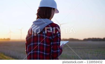 Female engineer taking notes with a tablet computer on a field with wind turbines, as the sun sets. Concept of clean energy and engineering audit 118950432