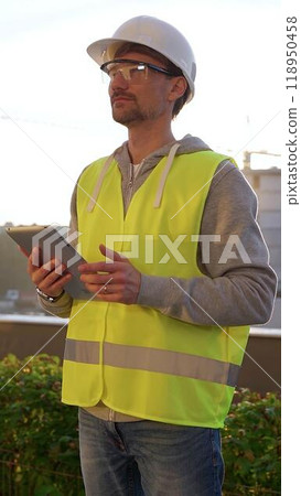 Man engineer with white helmet and safety vest is taking notes on a digital tablet while inspecting a construction site at sunrise in early morning, vertical front view. Architecture concept 118950458