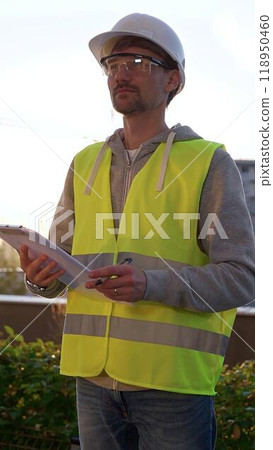Man engineer with white helmet and safety vest is taking notes on a clipboard while inspecting a construction site at sunrise in early morning, vertical front view. Architecture and engineering 118950460
