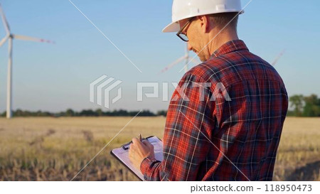 Man engineer at work is wearing a white protective helmet and taking notes with a clipboard in a field with wind turbines, as the sun sets. Clean energy concept 118950473