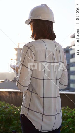 Female construction engineer wearing white checked blouse and hard hat is using tablet computer while inspecting a building site at sunset, back and vertical view. Architecture concept Female construction engineer wearing white checked blouse and hard hat is using tablet computer while inspecting a building site at sunset, back and vertical view. Architecture concept 118950509