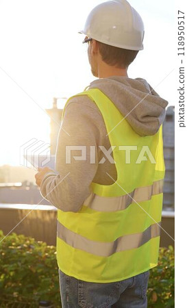 Man an architect or engineer with white helmet and safety vest is taking notes on a digital tablet while inspecting a construction site at sunrise, vertical back view. Architecture concept Man an architect or engineer with white helmet and safety vest is taking notes on a digital tablet while inspecting a construction site at sunrise, vertical back view. Architecture concept 118950517