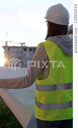 Woman an architect or engineer wearing safety hard hat and vest holding blueprint while inspecting a construction site at sunrise, vertical back view. Architecture and engineering 118950518