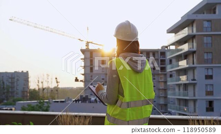 Construction female engineer taking notes and writing on clipboard while inspecting a building site at sunset. Industry development concept Construction female engineer taking notes and writing on clipboard while inspecting a building site at sunset. Industry development concept 118950589