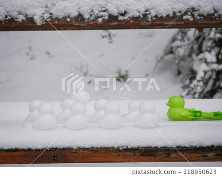 Snowmen in the shape of ducks. Ducks made of snow stand on a park bench next to a device for making snowballs 118950623