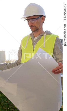 Man an architect or engineer wearing safety hard hat and vest holding blueprint and looking to construction site at sunrise, front vertical view. Architecture concept Man an architect or engineer wearing safety hard hat and vest holding blueprint and looking to construction site at sunrise, front vertical view. Architecture concept 118950637