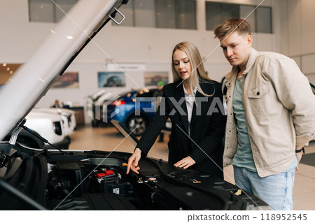 Pensive male customer in casual clothes standing thinking about buying car. Professional female salesperson with client in car showroom near open hood. Confident saleswoman explaining all car features 118952545