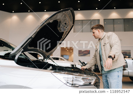 Serious pensive customer male looking inside opened car hood of new white automobile. Consumer young man in car dealership choosing auto. Concept of examining and buying new auto at showroom. 118952547