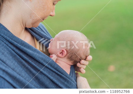 Mom walks with her newborn baby in a sling. This moment highlights the close bond between mother and child, promoting comfort, security, and the benefits of babywearing in everyday life Mom walks with her newborn baby in a sling. This moment highlights the close bond between mother and child, promoting comfort, security, and the benefits of babywearing in everyday life 118952766