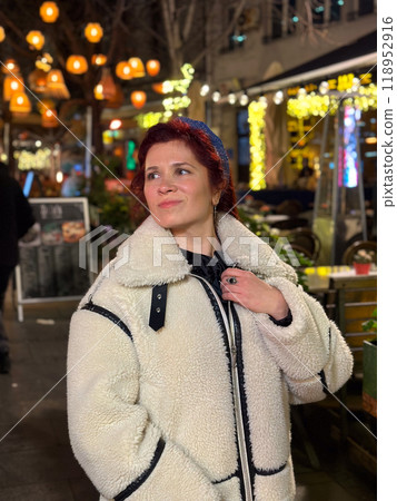 Millennial woman standing outdoors at Christmas night, wrapped in a cozy shearling jacket. the city lights in the background create a warm and Christmas holidays atmosphere, enhancing the peaceful and 118952916