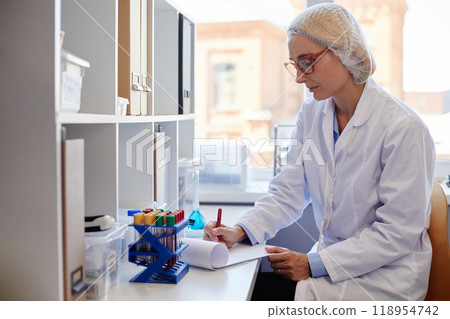 Side view of concentrated female chemist wearing lab coat and hairnet making records of blood test results in notepad while sitting at working table in laboratory, copy space Side view of concentrated female chemist wearing lab coat and hairnet making records of blood test results in notepad while sitting at working table in laboratory, copy space 118954742