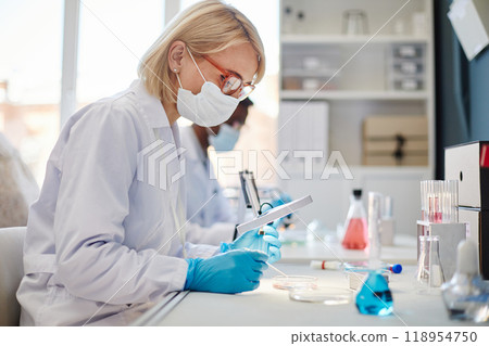SIde view of female biochemist in protective mask looking through magnifying glass at bacteria on petri dish studying microorganisms while working with pipette at table in laboratory, copy space 118954750