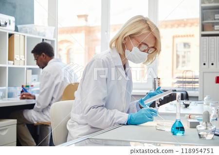 SIde view of female biochemist in protective mask studying bacteria in petri dish working with pipette while sitting at table in laboratory of scientific research service, copy space SIde view of female biochemist in protective mask studying bacteria in petri dish working with pipette while sitting at table in laboratory of scientific research service, copy space 118954751