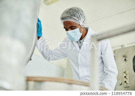 Medium shot of African American male technician in protective mask opening lid of huge steel tank inspecting production process on shop floor of pharmaceutical manufacturing plant, copy space Medium shot of African American male technician in protective mask opening lid of huge steel tank inspecting production process on shop floor of pharmaceutical manufacturing plant, copy space 118954778